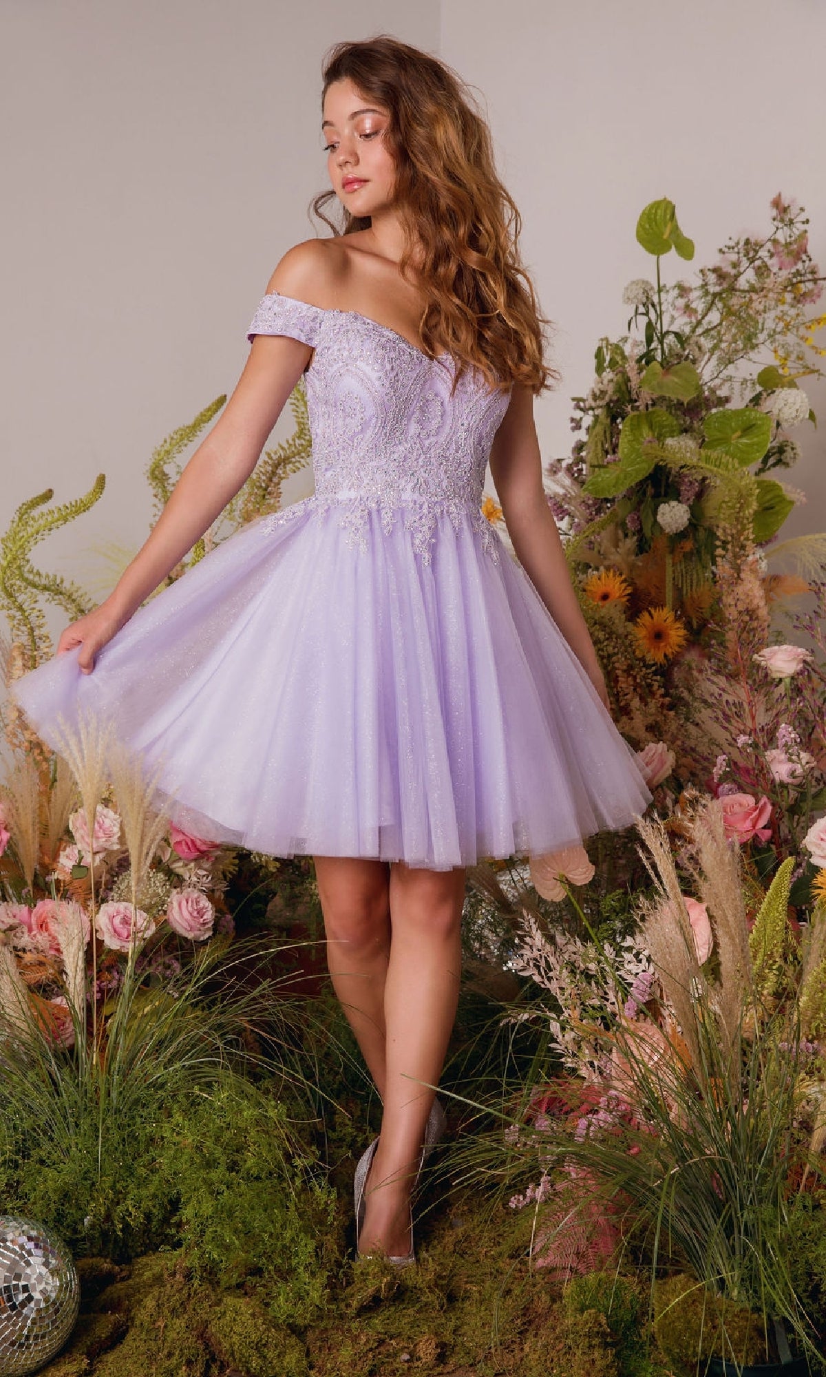 A woman wearing the Eureka 9833 Off-the-Shoulder Short Hoco Dress in lavender stands among assorted flowers and greenery, gazing to the side.